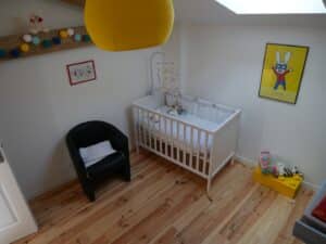 Nursery lumineuse avec lit bébé blanc, fauteuil noir, lampe jaune. Sol bois, affiche T'choupi fait des bêtises et déco colorée.