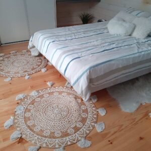 Chambre cosy: lit rayé bleu/gris, coussins blancs. Deux tapis ronds en jute bohème à motifs sur sol bois clair. Décor mural cœurs.
