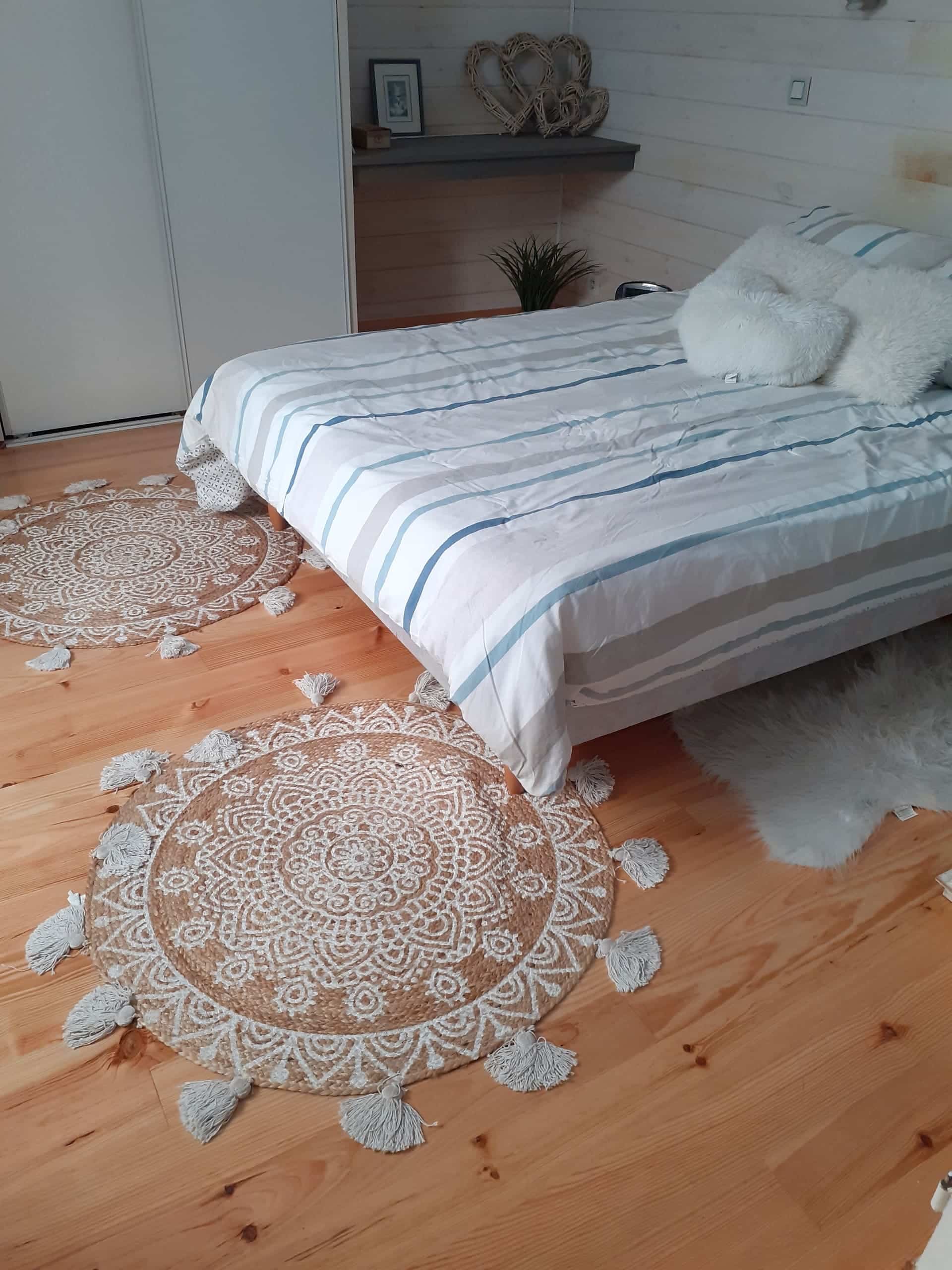 Chambre cosy: lit rayé bleu/gris, coussins blancs. Deux tapis ronds en jute bohème à motifs sur sol bois clair. Décor mural cœurs.