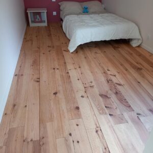 Chambre enfant lumineuse avec parquet en pin clair, lit simple blanc, et mur rose. Une table de chevet et une peluche bleue.