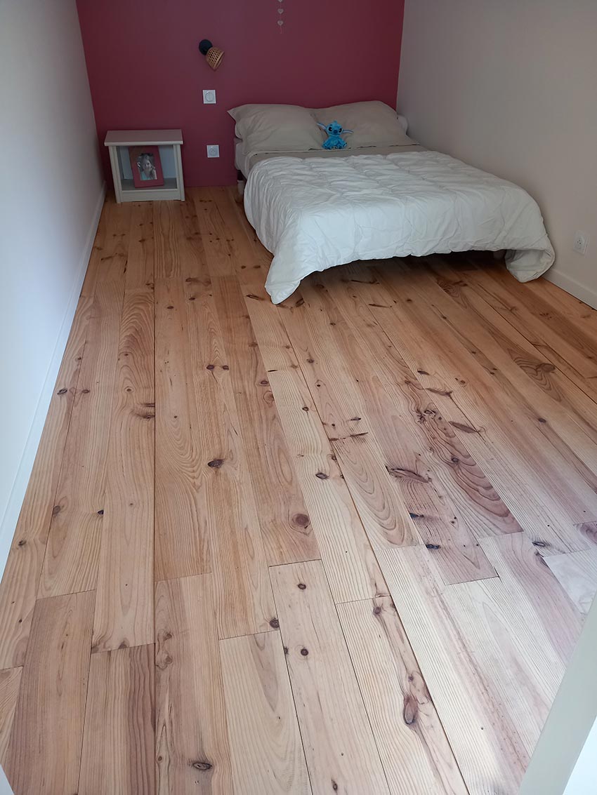 Chambre enfant lumineuse avec parquet en pin clair, lit simple blanc, et mur rose. Une table de chevet et une peluche bleue.