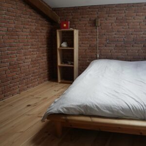 Chambre mansardée avec murs en briques rouges et parquet bois clair. Lit en bois, couette rayée, étagère, poutre apparente.