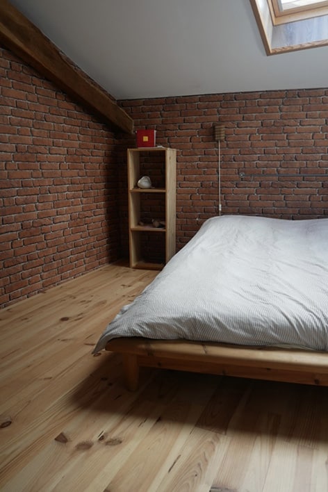 Chambre mansardée avec murs en briques rouges et parquet bois clair. Lit en bois, couette rayée, étagère, poutre apparente.