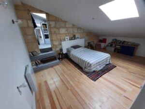 Chambre mansardée lumineuse avec lit rayé, parquet clair et mur en pierre. Escalier vers une pièce en mezzanine.