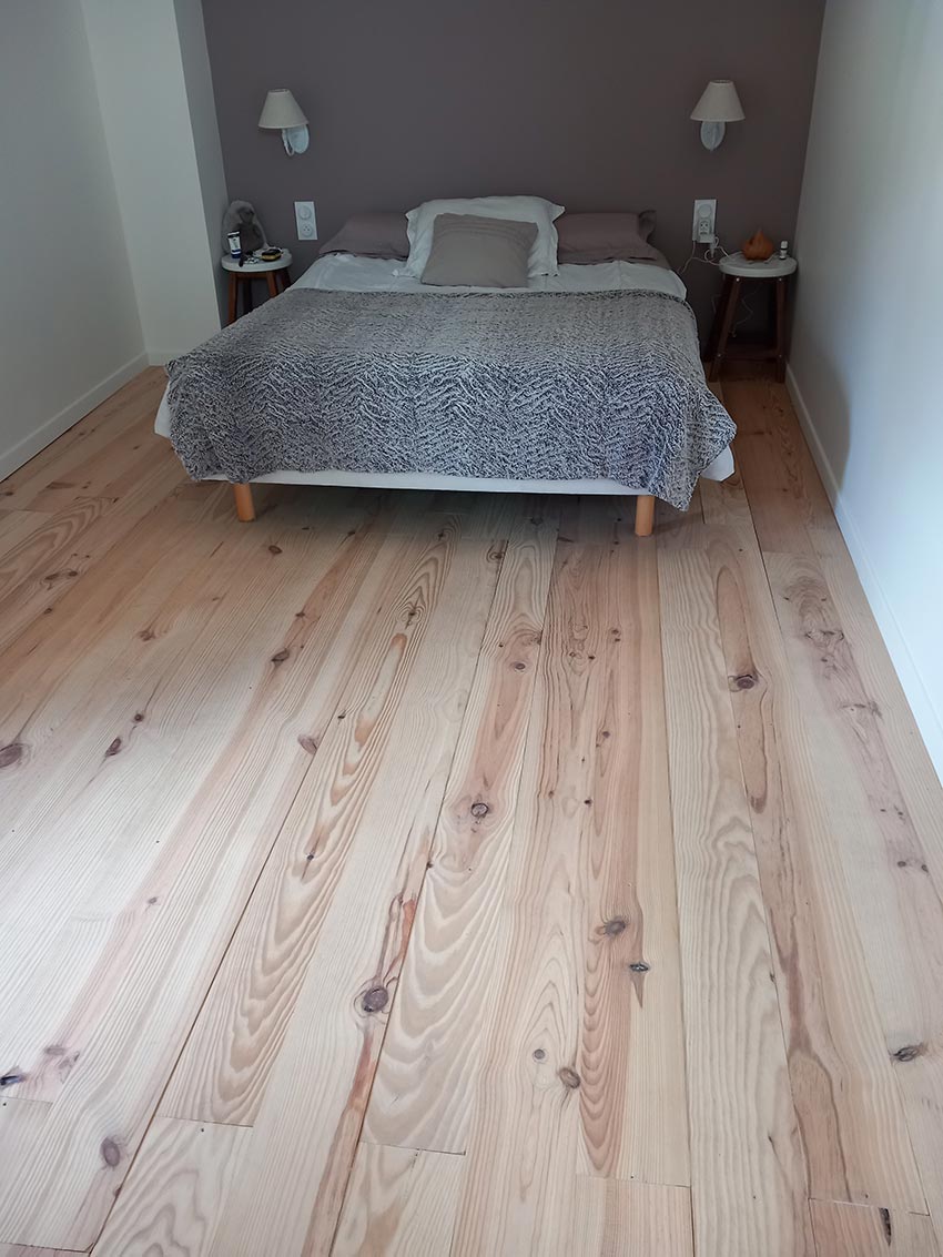 Chambre élégante avec grand lit gris sur parquet clair en bois, murs gris et blancs. Deux tables de chevet avec lampes.