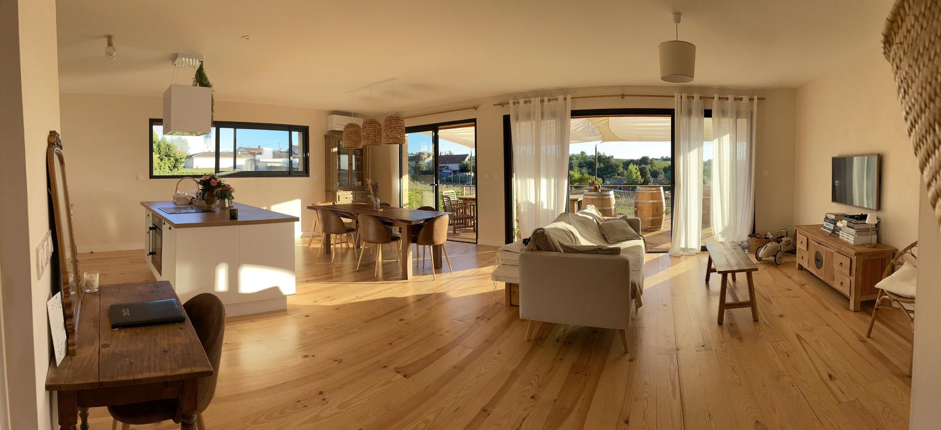 Grand espace de vie lumineux avec cuisine ouverte, salle à manger bois et salon. Terrasse avec vue sur vignoble et campagne ensoleillée.