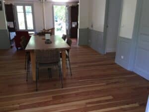 Grande pièce lumineuse avec parquet en bois clair, murs gris-blanc. Salle à manger équipée d'une table en bois massif et chaises en osier. Espace salon avec fauteuil rouge visible au fond.