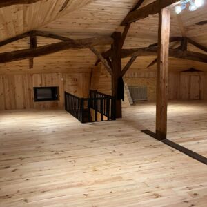 Grand loft en bois avec plafond cathédrale et poutres apparentes. Sol clair, murs lambrissés, escalier noir, fenêtre encastrée et éclairage moderne.
