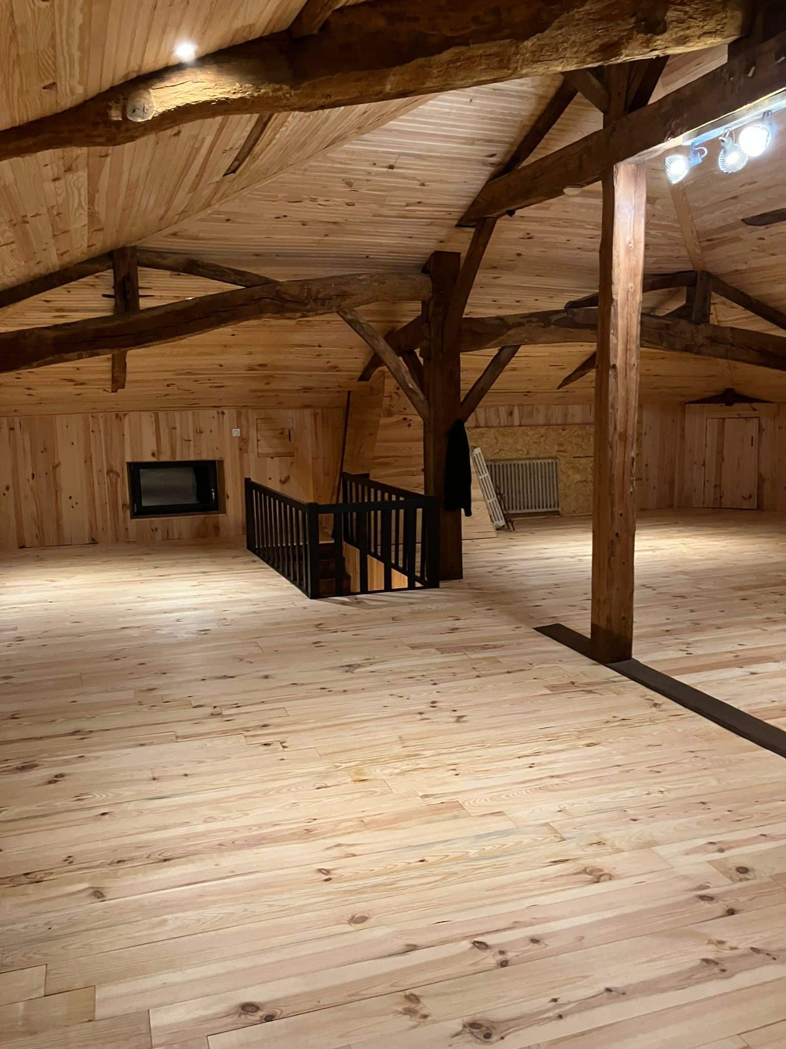 Grand loft en bois avec plafond cathédrale et poutres apparentes. Sol clair, murs lambrissés, escalier noir, fenêtre encastrée et éclairage moderne.