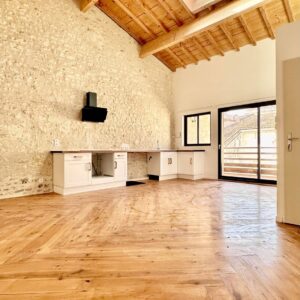 Loft rénové avec mur en pierres apparentes, plafond et sol en bois clair. Cuisine nue et grande baie vitrée offrant vue sur toits.