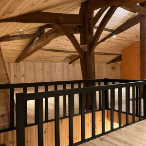 Mezzanine en bois clair avec plafond lambrissé et poutres apparentes. Rambarde noire moderne au-dessus d'un escalier.
