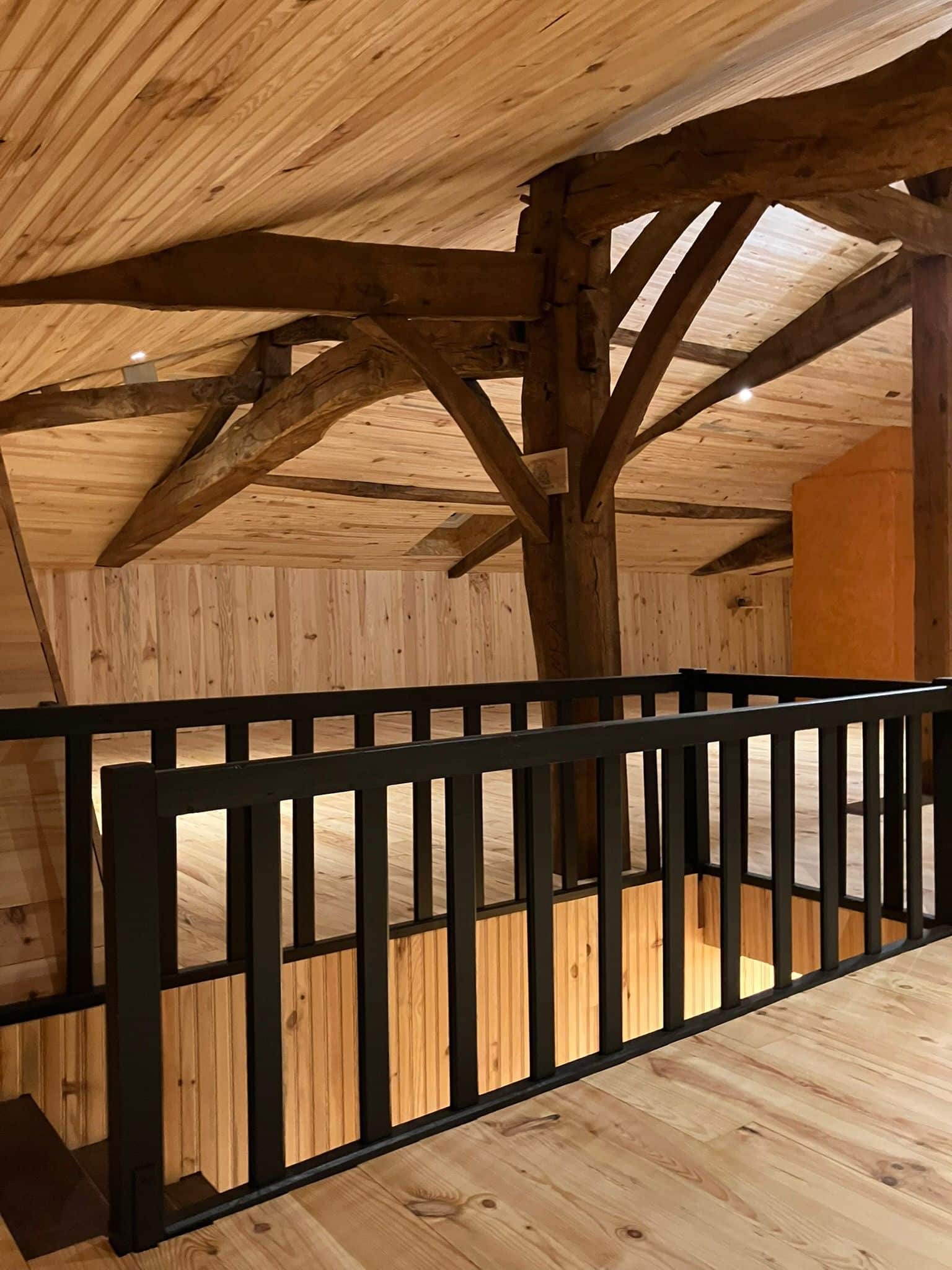 Mezzanine en bois clair avec plafond lambrissé et poutres apparentes. Rambarde noire moderne au-dessus d'un escalier.