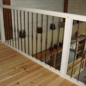 Mezzanine avec plancher bois clair, vue sur salon-cuisine à travers un garde-corps unique avec balustres en métal foncé et tasses décoratives.