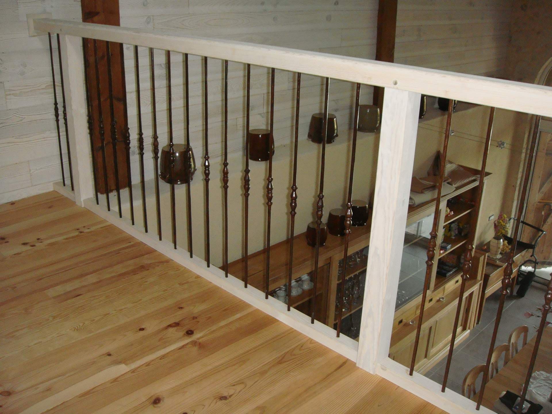 Mezzanine avec plancher bois clair, vue sur salon-cuisine à travers un garde-corps unique avec balustres en métal foncé et tasses décoratives.