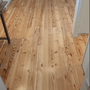 Parquet en bois clair fraîchement posé avec veinures visibles, dans une pièce aux murs blancs, près d'un établi et d'un cadre de porte.