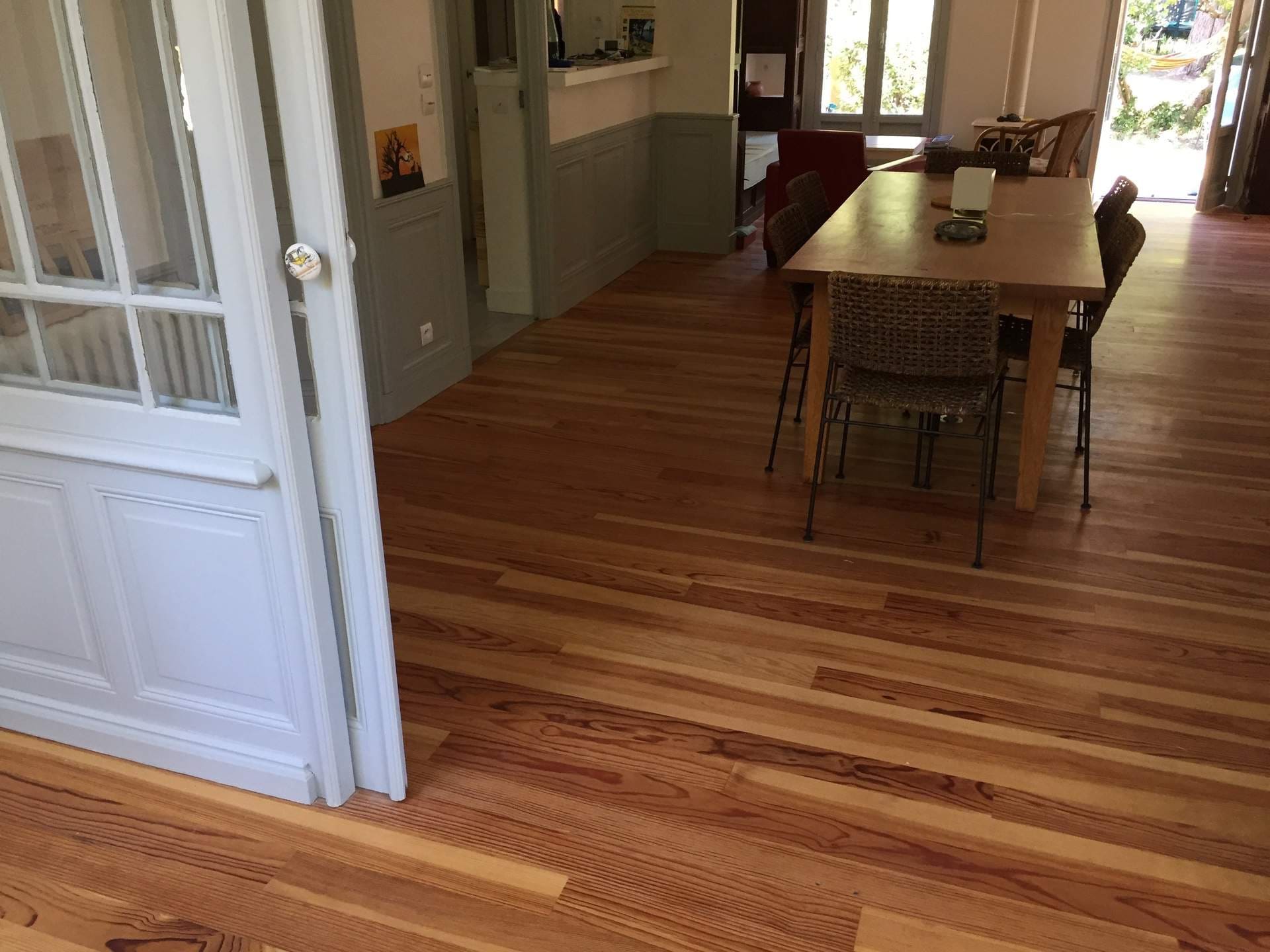 Salle lumineuse avec parquet en bois. Porte française blanche, table et chaises en osier. Grandes fenêtres donnent sur le jardin.