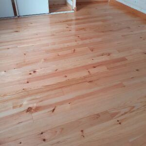 Parquet en bois clair avec nœuds et grain apparents dans une pièce lumineuse. Armoire blanche coulissante à gauche, mur lambrissé et prises à droite.