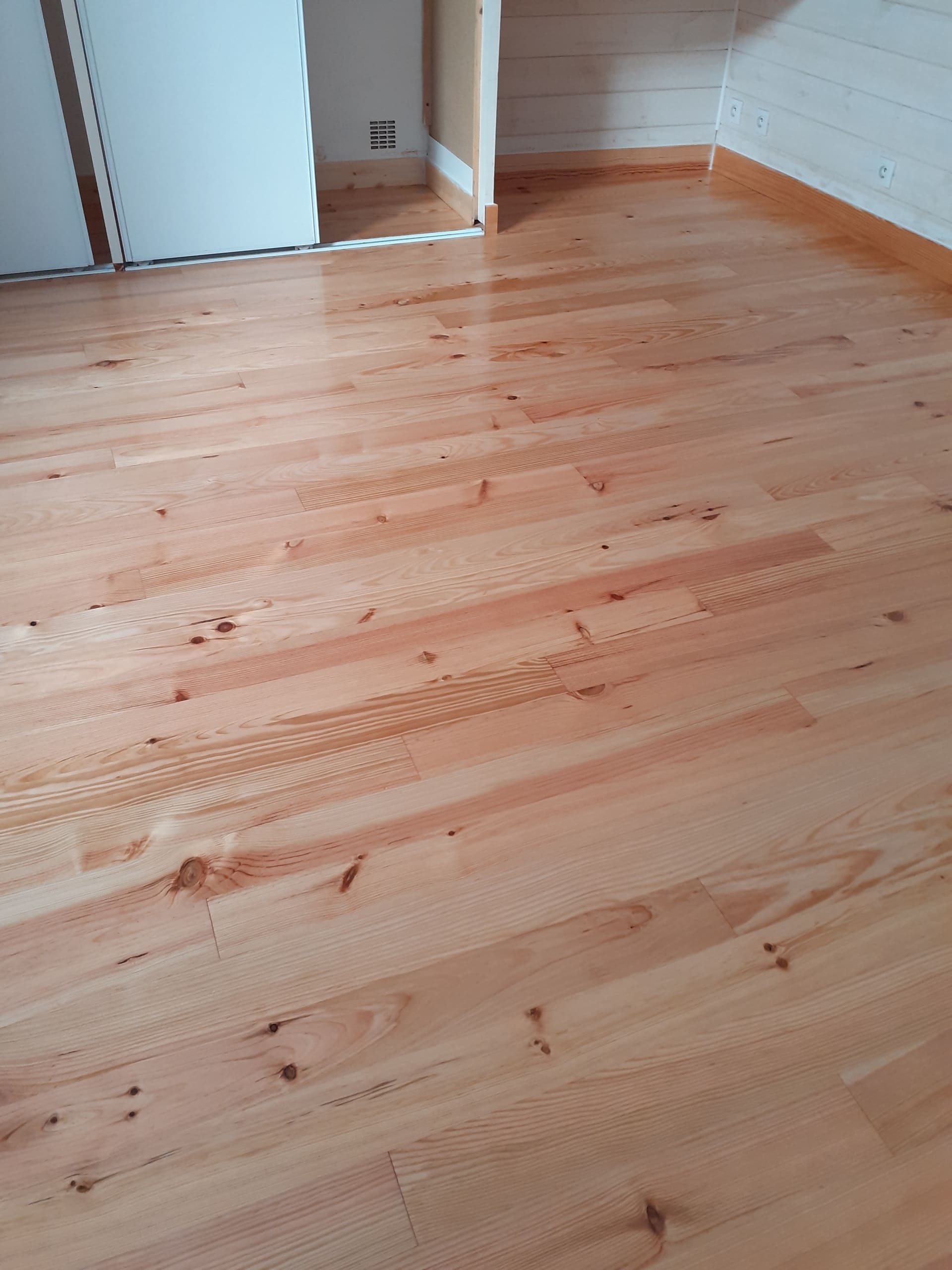 Parquet en bois clair avec nœuds et grain apparents dans une pièce lumineuse. Armoire blanche coulissante à gauche, mur lambrissé et prises à droite.