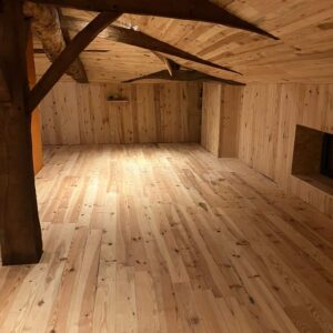 Pièce entièrement boisée avec parquet clair, murs lambrissés et plafond à poutres sombres. Un grand tronc d'arbre brut supporte la structure.