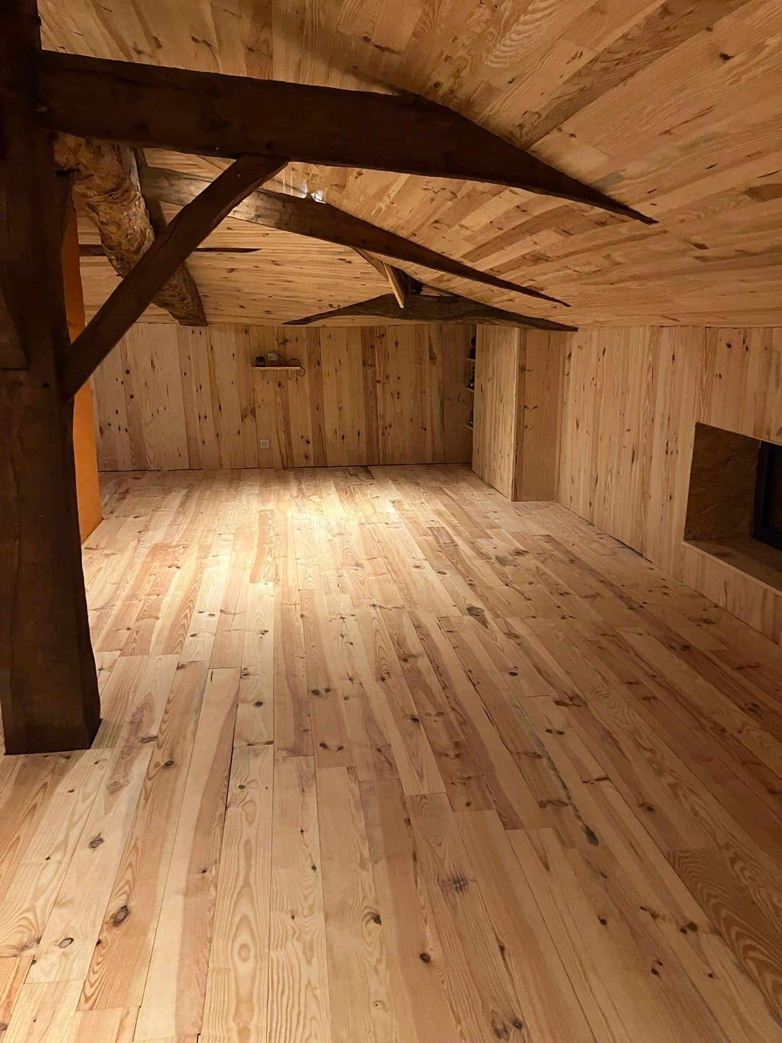Pièce entièrement boisée avec parquet clair, murs lambrissés et plafond à poutres sombres. Un grand tronc d'arbre brut supporte la structure.