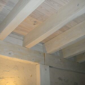 Plafond en bois clair avec poutres apparentes et planches de pin, soutenu par une charpente et un mur texturé ocre.