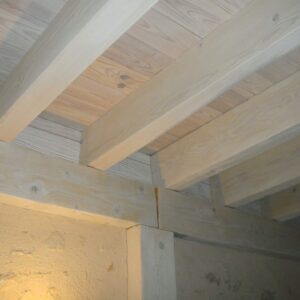 Plafond en bois clair avec poutres apparentes et mur texturé. Une colonne en bois soutient la structure, éclairée par une lumière chaude.