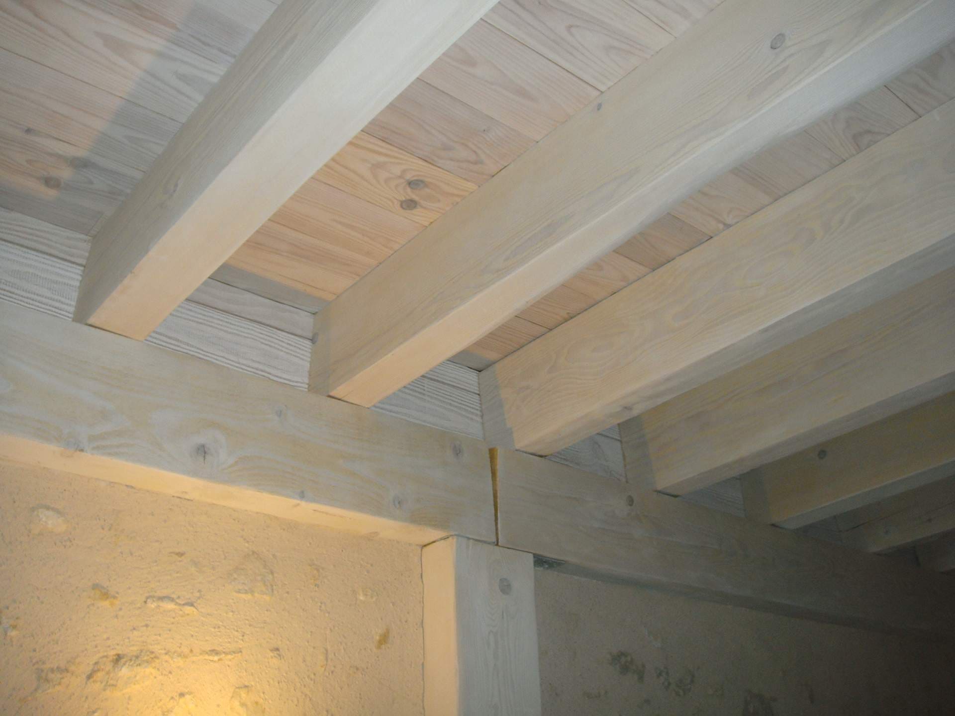 Plafond en bois clair avec poutres apparentes et mur texturé. Une colonne en bois soutient la structure, éclairée par une lumière chaude.