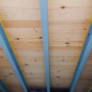 Plafond moderne avec des poutres bleues et un cadre jaune supportant des planches de bois clair. Structure architecturale distinctive.