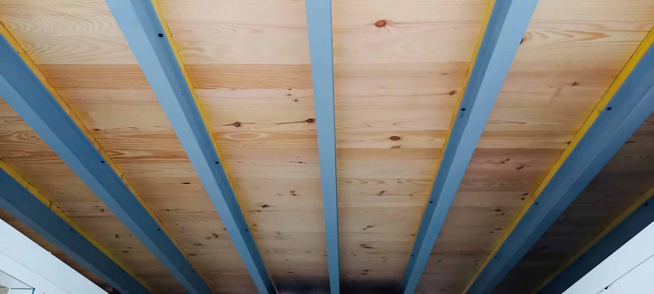 Plafond moderne avec des poutres bleues et un cadre jaune supportant des planches de bois clair. Structure architecturale distinctive.
