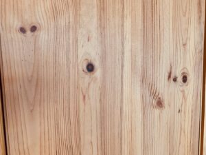 Panneau en bois clair avec des veines verticales et plusieurs nœuds distincts, dont certains ressemblent à des yeux.