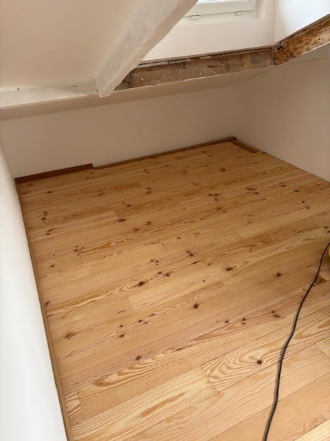 Plancher en bois clair neuf posé dans une pièce aux combles aménagés, avec lames de pin et poutre apparente.