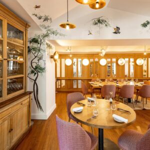 Élégante salle à manger de restaurant. Muraux d'oiseaux et branches, grand buffet en bois, tables rondes avec chaises roses, lumière dorée.