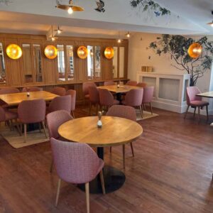 Salle de restaurant chaleureuse : tables en bois, chaises roses, boiseries claires, décor mural d'arbres/oiseaux et lumières dorées.