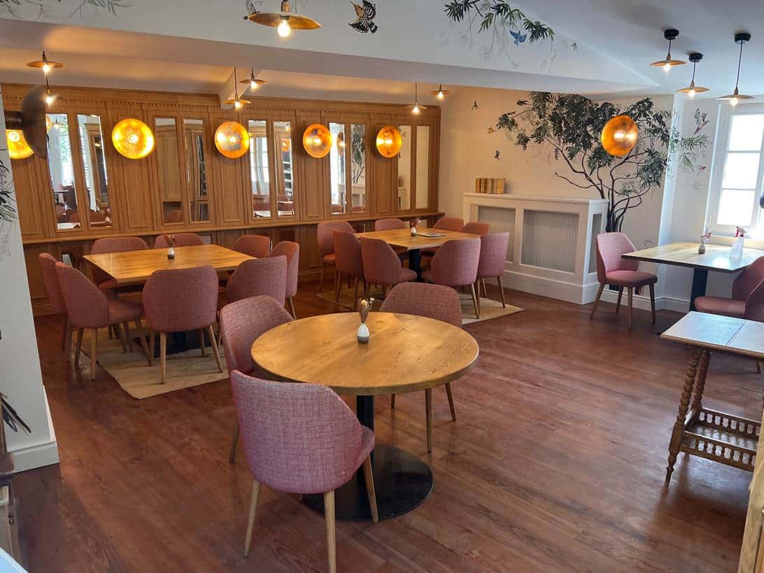 Salle de restaurant chaleureuse : tables en bois, chaises roses, boiseries claires, décor mural d'arbres/oiseaux et lumières dorées.