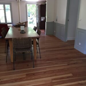 Salle à manger lumineuse avec grande table en bois et chaises en osier. Parquet clair, fenêtres ouvertes sur l'extérieur et boiseries grises élégantes.
