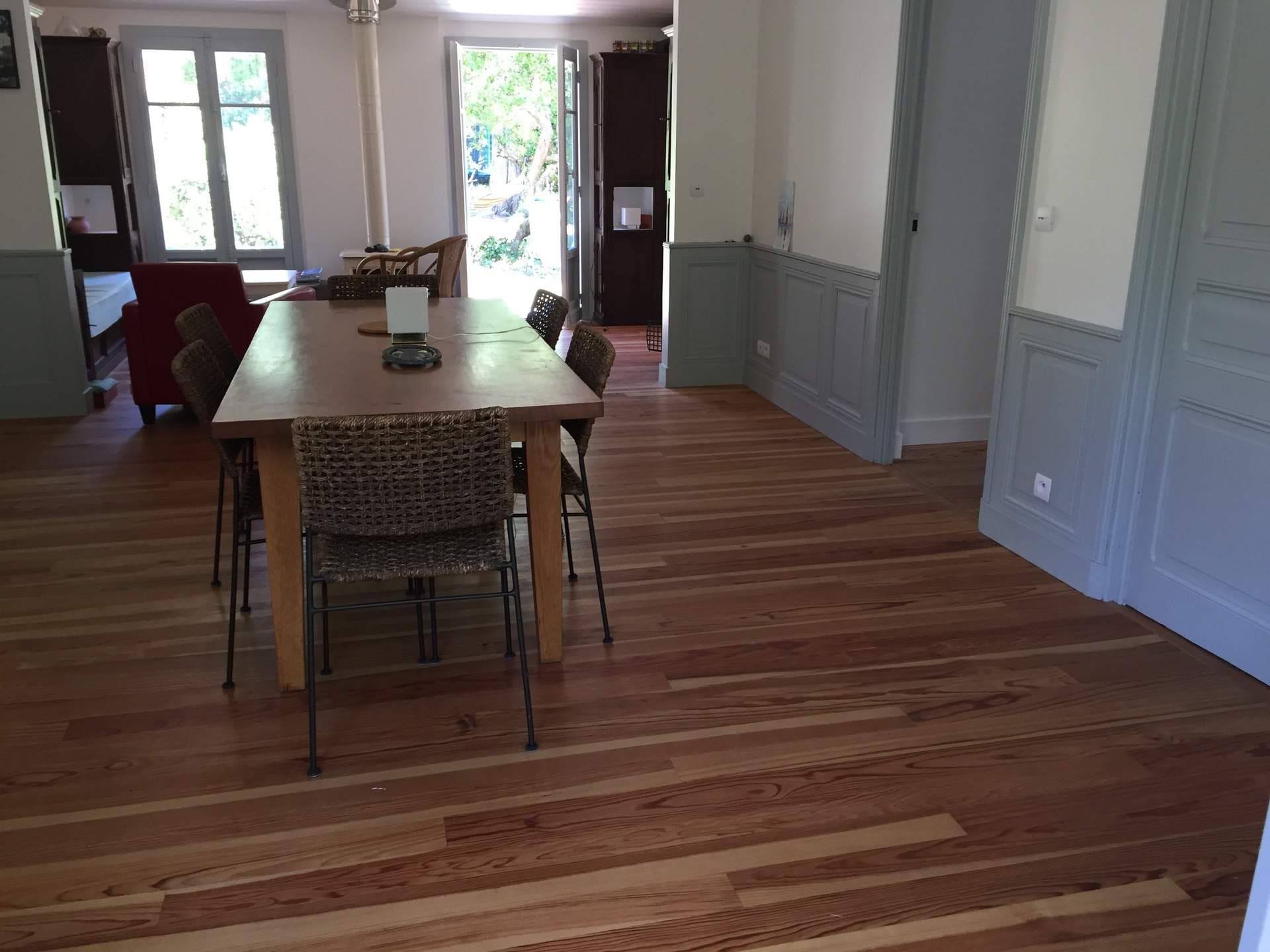 Salle à manger lumineuse avec grande table en bois et chaises en osier. Parquet clair, fenêtres ouvertes sur l'extérieur et boiseries grises élégantes.