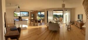 Salon, cuisine et salle à manger ouverts, lumineux avec sol bois clair. Vue sur terrasse, vignobles et fûts depuis de larges baies vitrées.