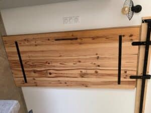 Tête de lit rustique en bois clair avec grain apparent et nœuds, fixée au mur. Supports en métal noir industriel sur fond blanc. Prises visibles.